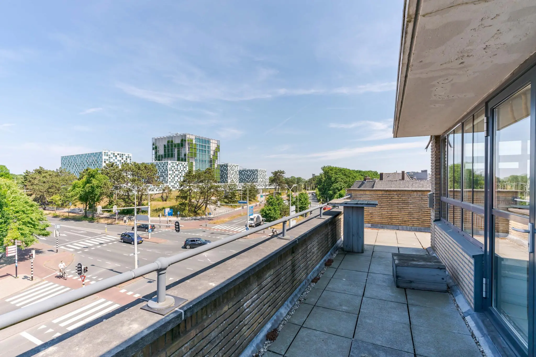 Uitzicht vanaf een balkon aan de Van Alkemadelaan op een kruispunt en moderne kantoorgebouwen te midden van groen.